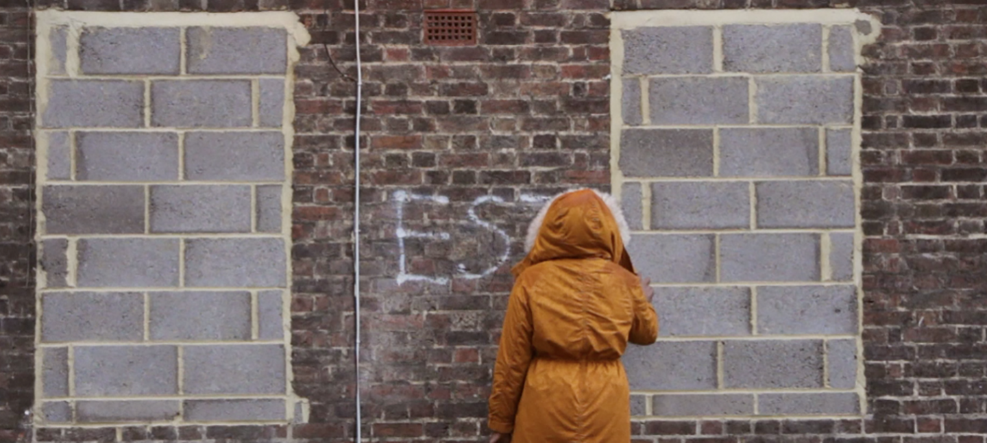 A still from 'Estate: a Reverie', 2015 by Andrea Luka Zimmerman. It shows someone in a orange coat with their hood up spraypainting a brick wall