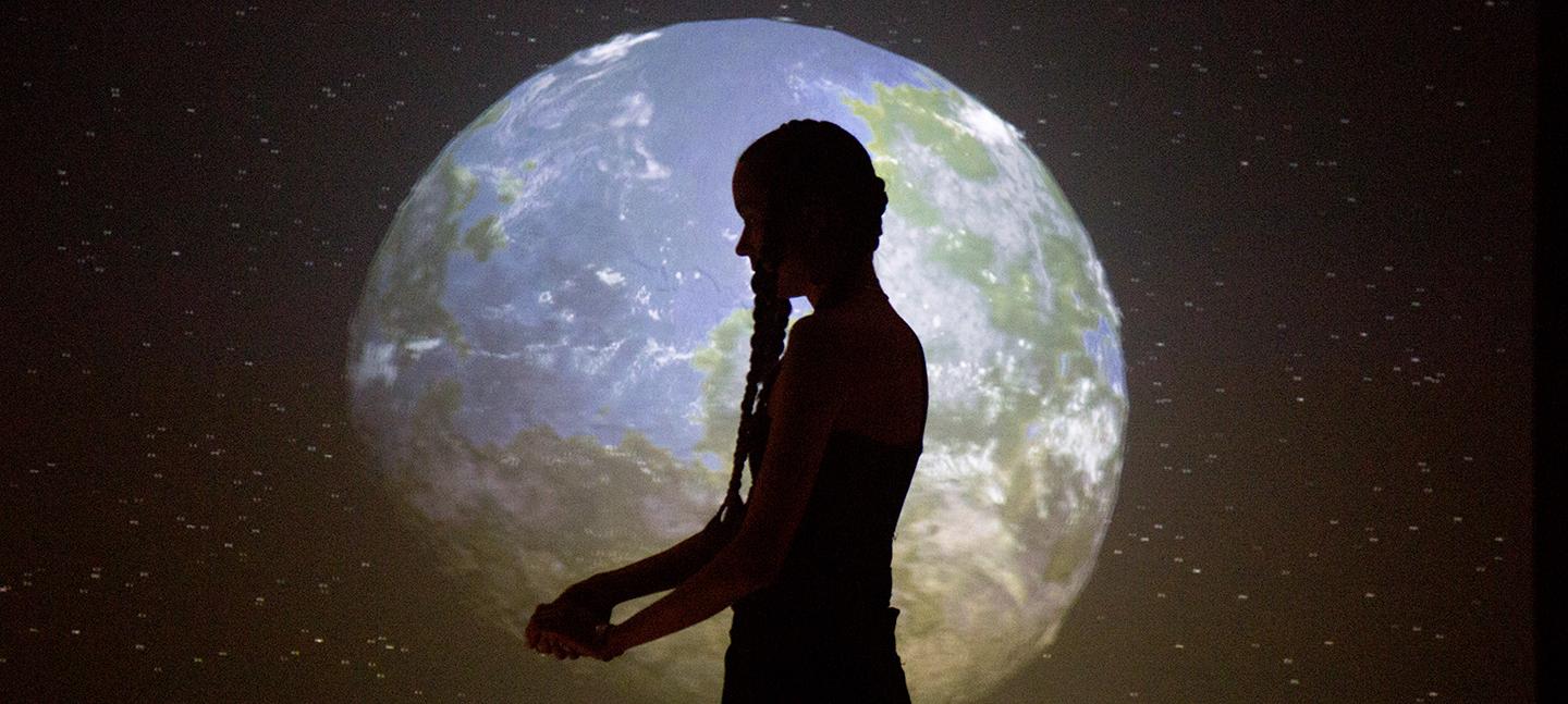 A woman (Anna Mikkola) standings in front of a projection of the world, as seen from space