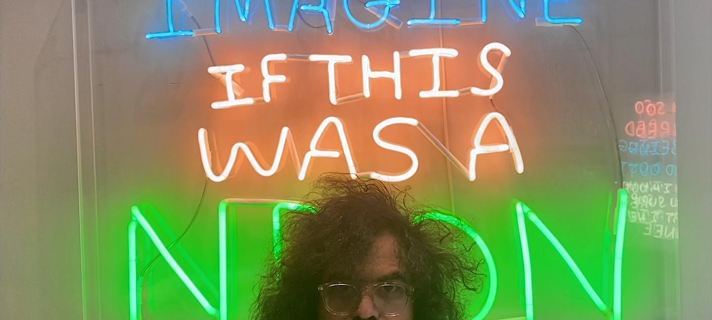 A photo of Babak Ganjei stood in front of one of his artworks. Babak stands at the front of the shot, and only his eyes and curly dark hair can be seen peering above. In the background is a neon artwork, which says 'Imagine if this were a neon'