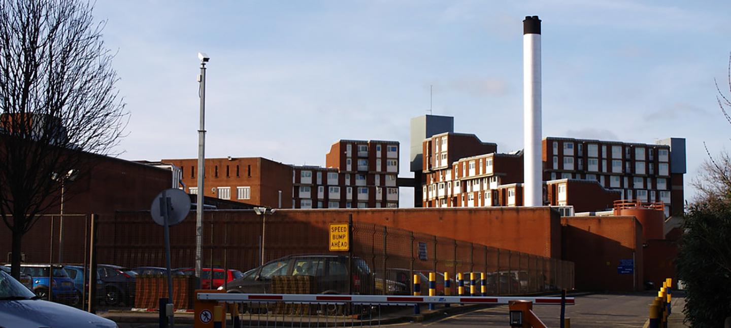 A photo of Holloway Prison. In the foreground you can see a gated carpark and a red and white barrier., alongside a CCTV pole. In the background Holloway Prison looms against a cold, blue, winter sky. A skeleton-like tree is to the left.