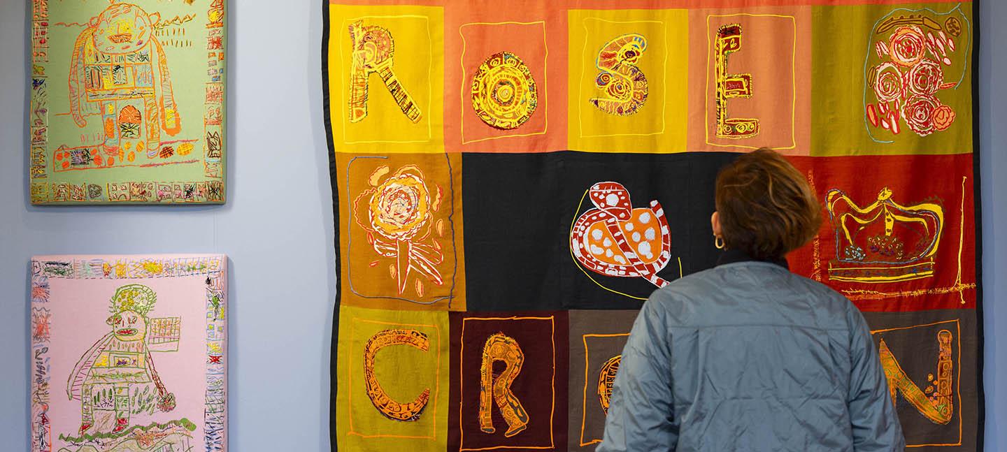 A woman stands in front of some colourful textile art hung up on the wall. The large patchwork tapestry reads ROSE CROWN.