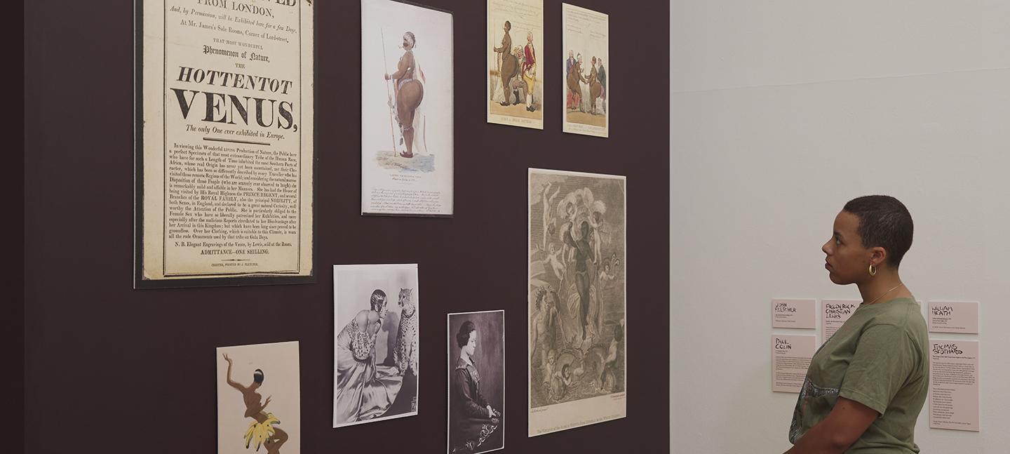 A young woman looks on at artworks carefully hung on a wall, painted a rich velvet brown.