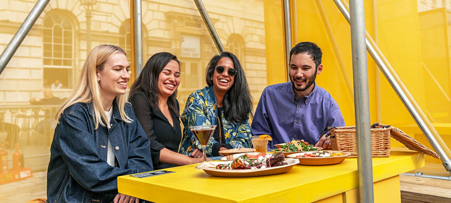 A group of four enjoying food and drink at a colourful booth designed by Yinka Ilori, located in the courtyard at Somerset House
