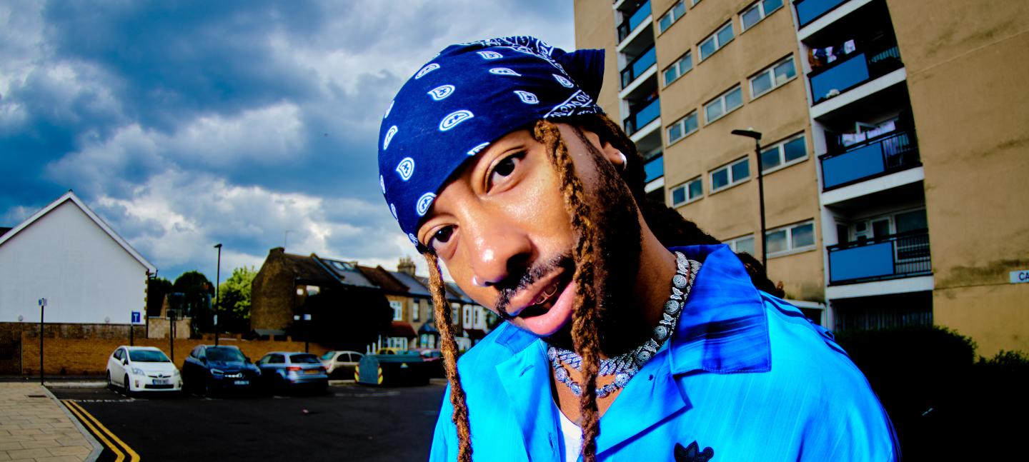 Donnie Sunshine wears a blue headscarf with two braids framing his face. They are wearing a blue adidas shirt.