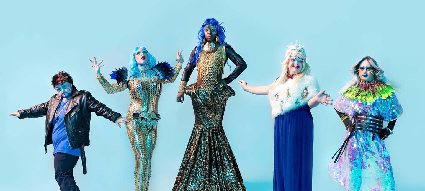 A photo of Drag Queens and Kings in stunning, sparkling outfits, each posing in an individual way. They pose against a turquoise background.