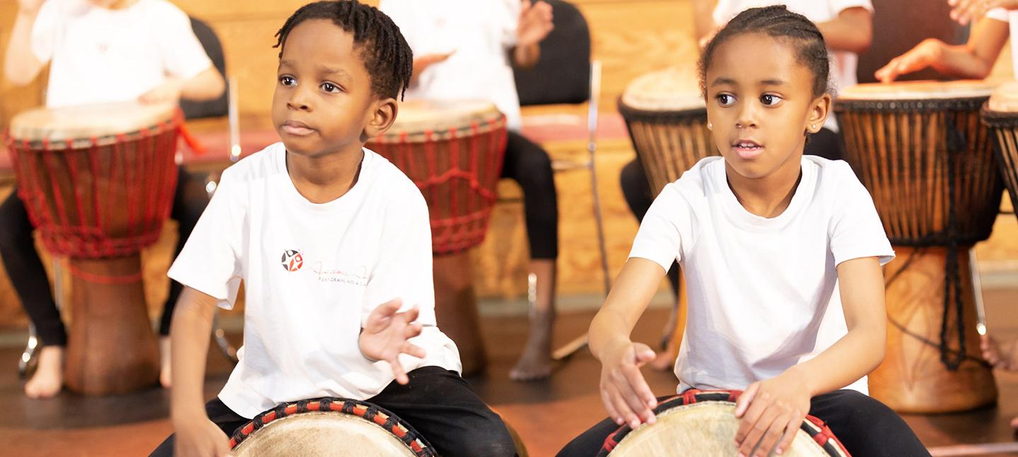 Two little children play drums at a workshop. They are concentrating and quietly smiling.