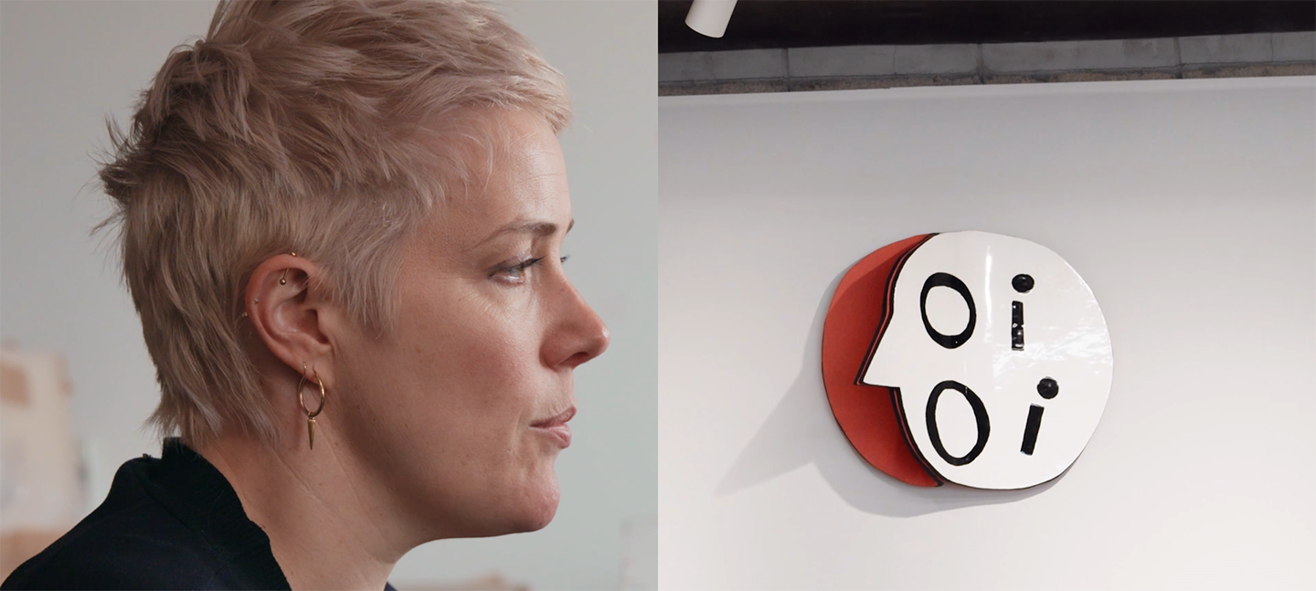 A film still from Emma Hart: Language and Class. On the left hand side is a profile view of the artist Emma Hart, a woman with short blonde hair, who is mid-speech. On the right-handside is a ceramic artwork that spells OI OI