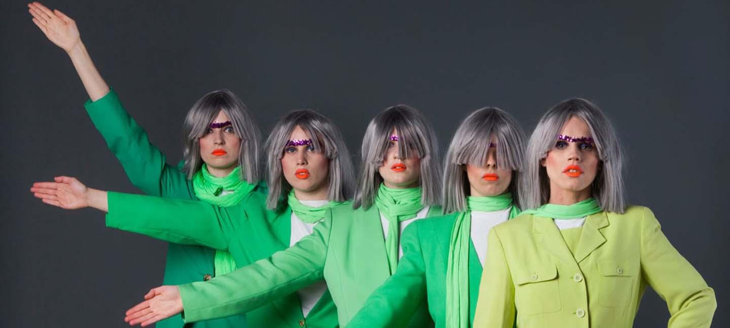 A photo of Figs in Wigs performers. Five people in grey, bob blunt cut wigs and green jackets and linen scarves around their necks stare into the camera. Each has their arm raised at a slightly different angle.