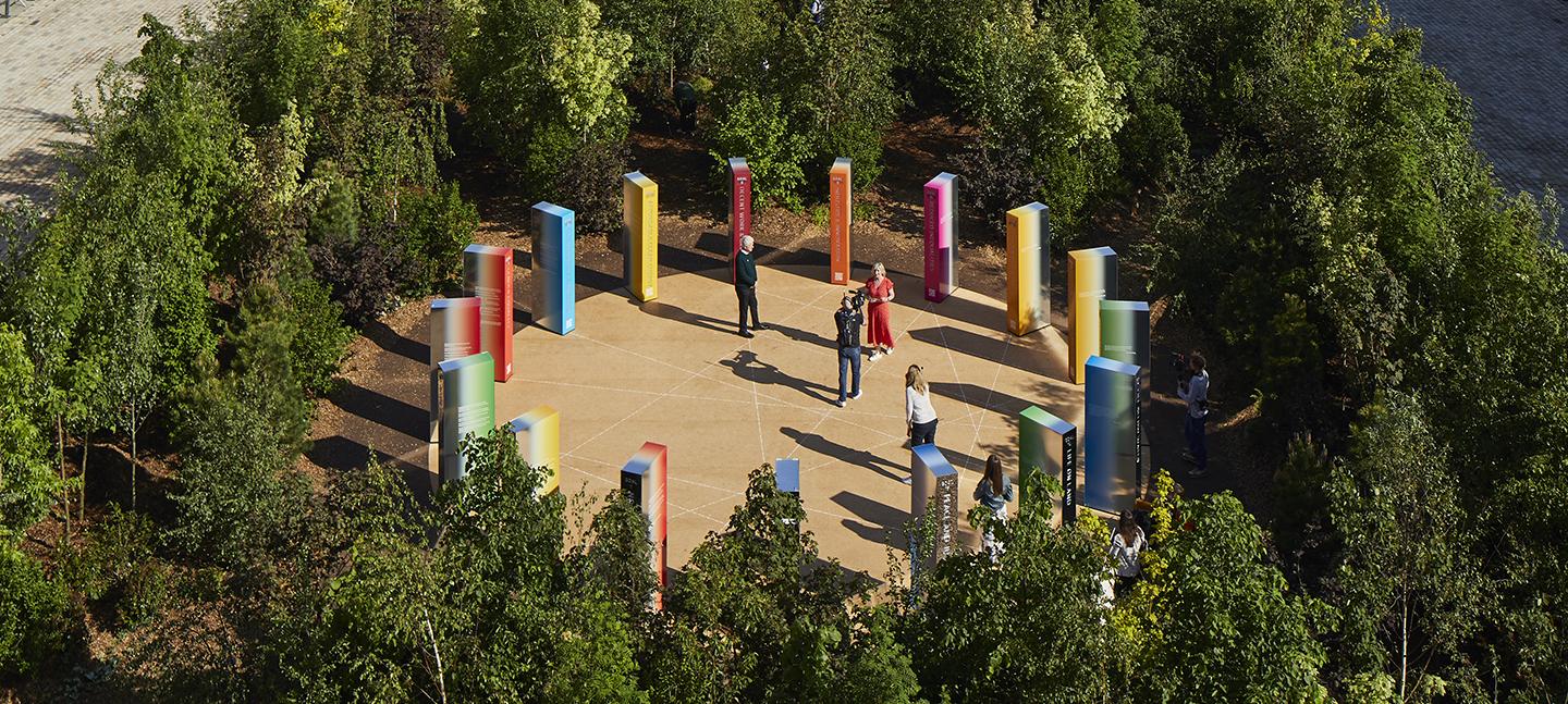 An aerial view of Forest for Change, focussing in on the installation at its centre. Lots of coloured obelisk shapes in a clearing, surrounded by a forest of trees.