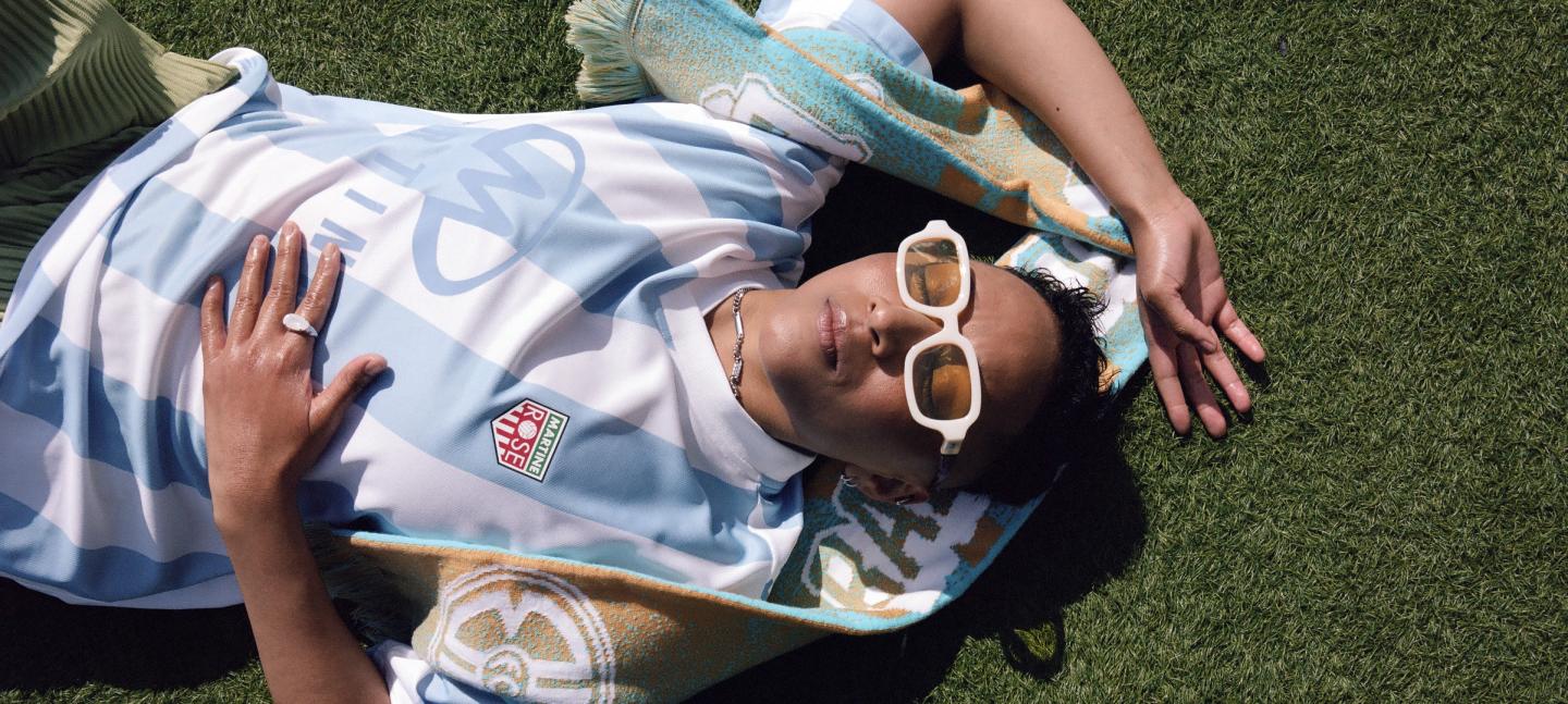GIN lays on the grass with the camera shooting them from above. They are bathed in sunshine and smiling, while wearing a football shirt and white sunglasses.