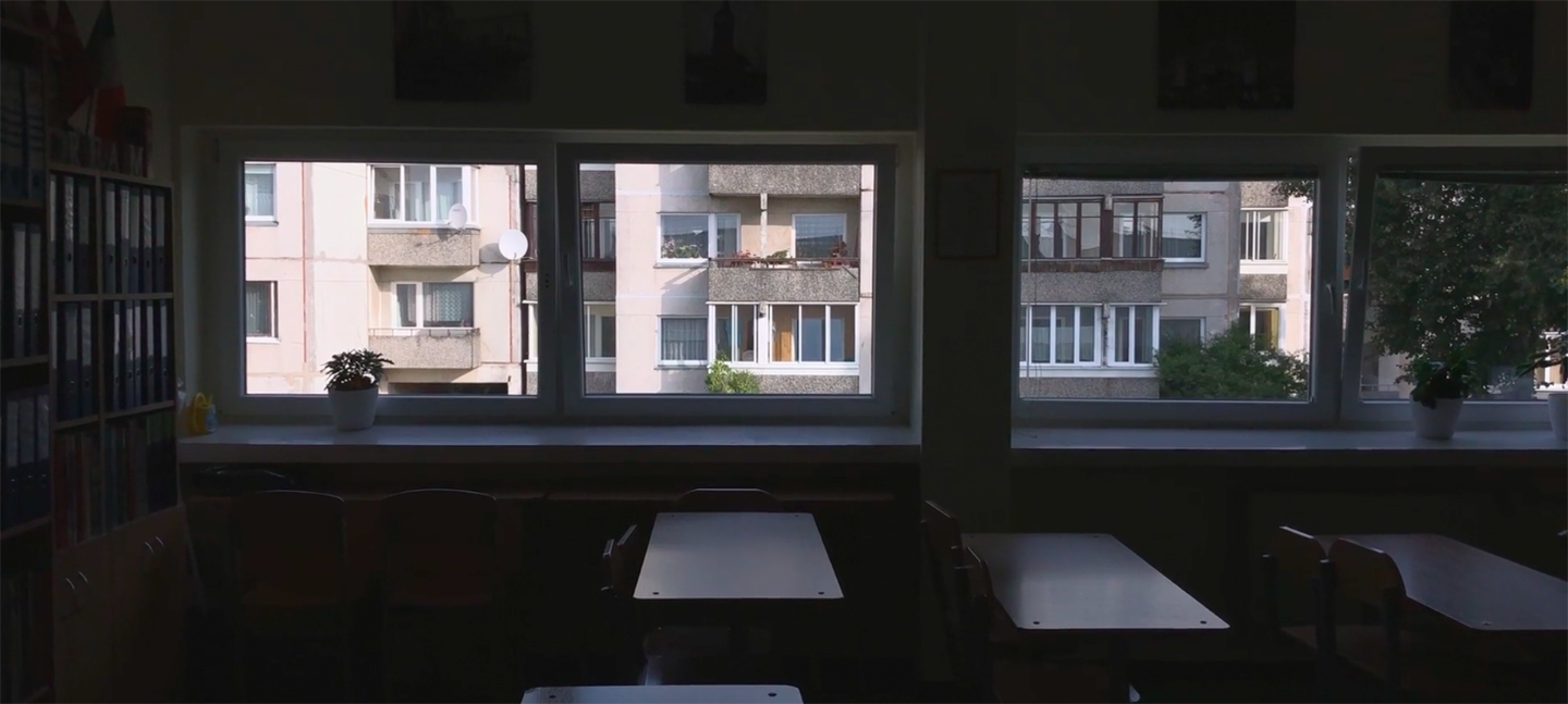 A still from Gerda Paliušytė's film Early Winter. It depicts a class room, with empty chairs and desks, with windows, looking out to buildings across the way.