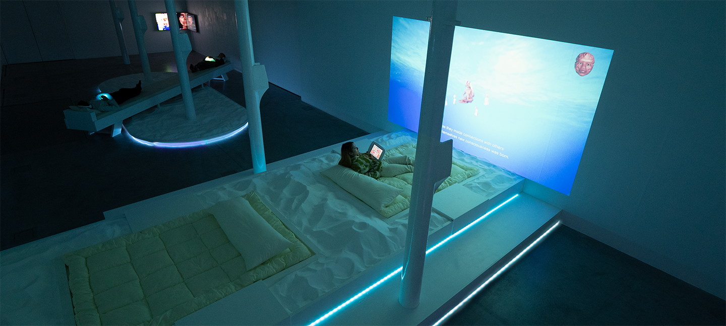 An installation image of an artwork by the collective Keiken. In a blue-lit gallery space are projections and TVs on the wall. On the floor are pillowy mattresses and cushions, surrounded by sand. People are reclining on these structures.