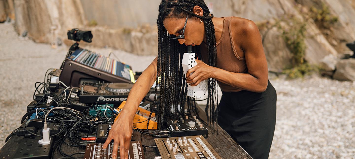 A photo of Nkisi performing on a beach at Nextones. Nkisi is shown in profile, with her decks and electronic sound equipment in front of her.