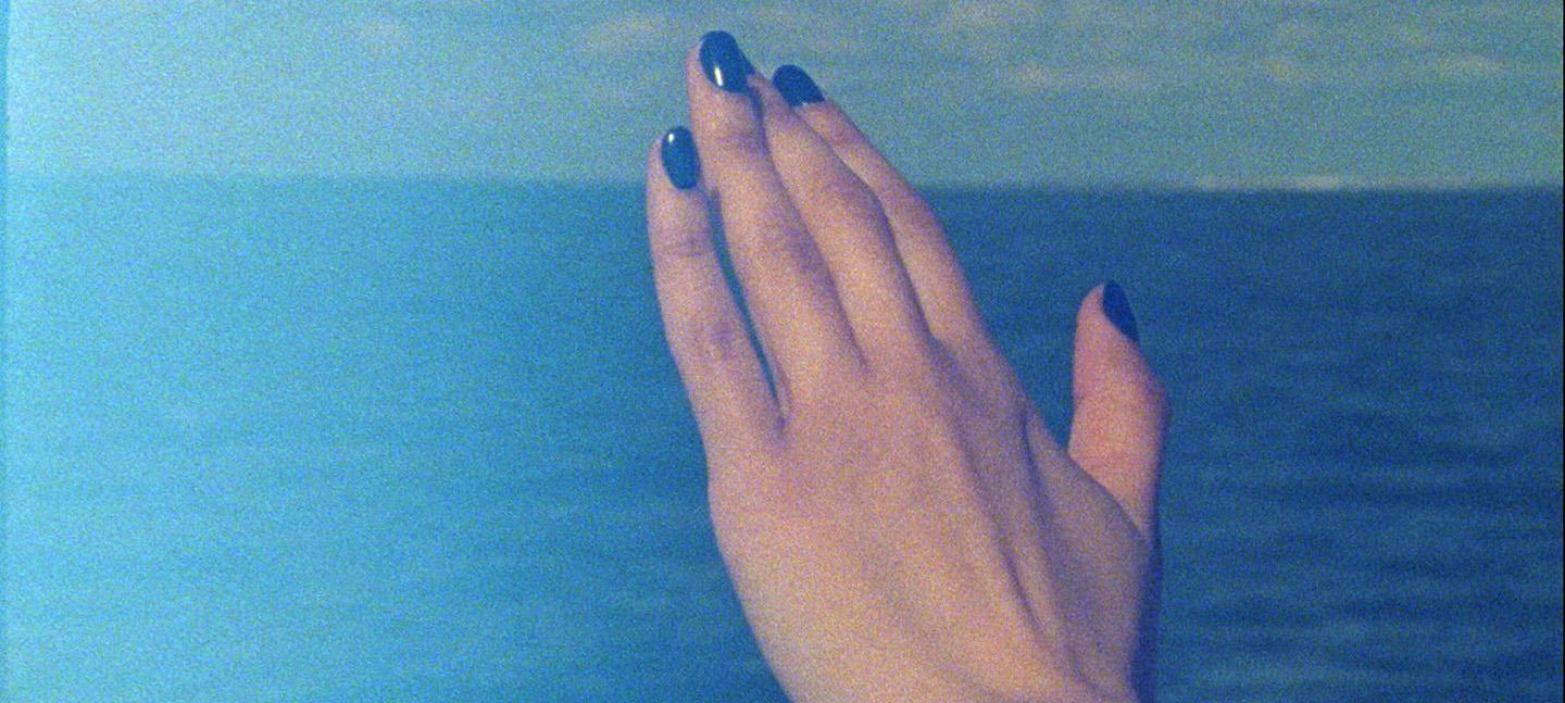 A still from Ana Vaz's film Occidente. It shows a woman's manicured hands with a shiny dark blue acrylic on a background of sea and sky,