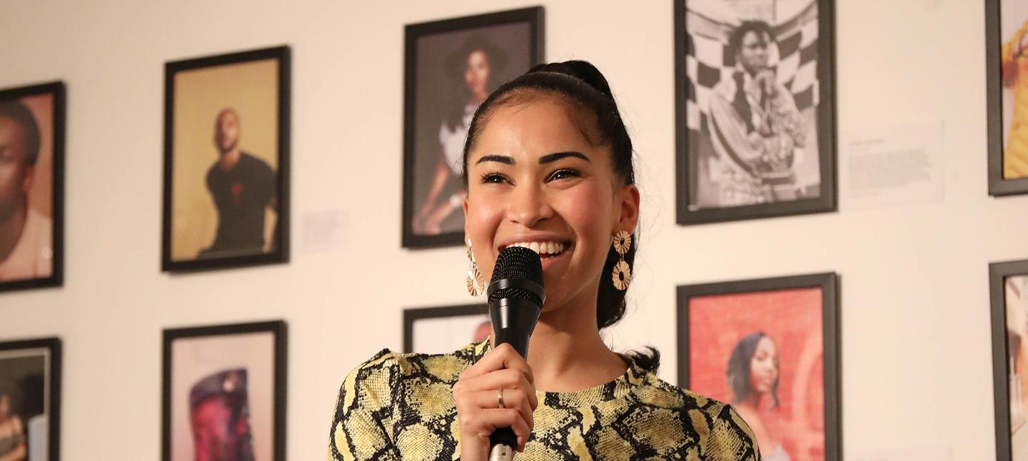 A photo of Swarzy smiling with a microphone in her hand. She stands in front of a wall of framed photos of Black creatives at an exhibition created by Swarzy, entitled Too Much Source.