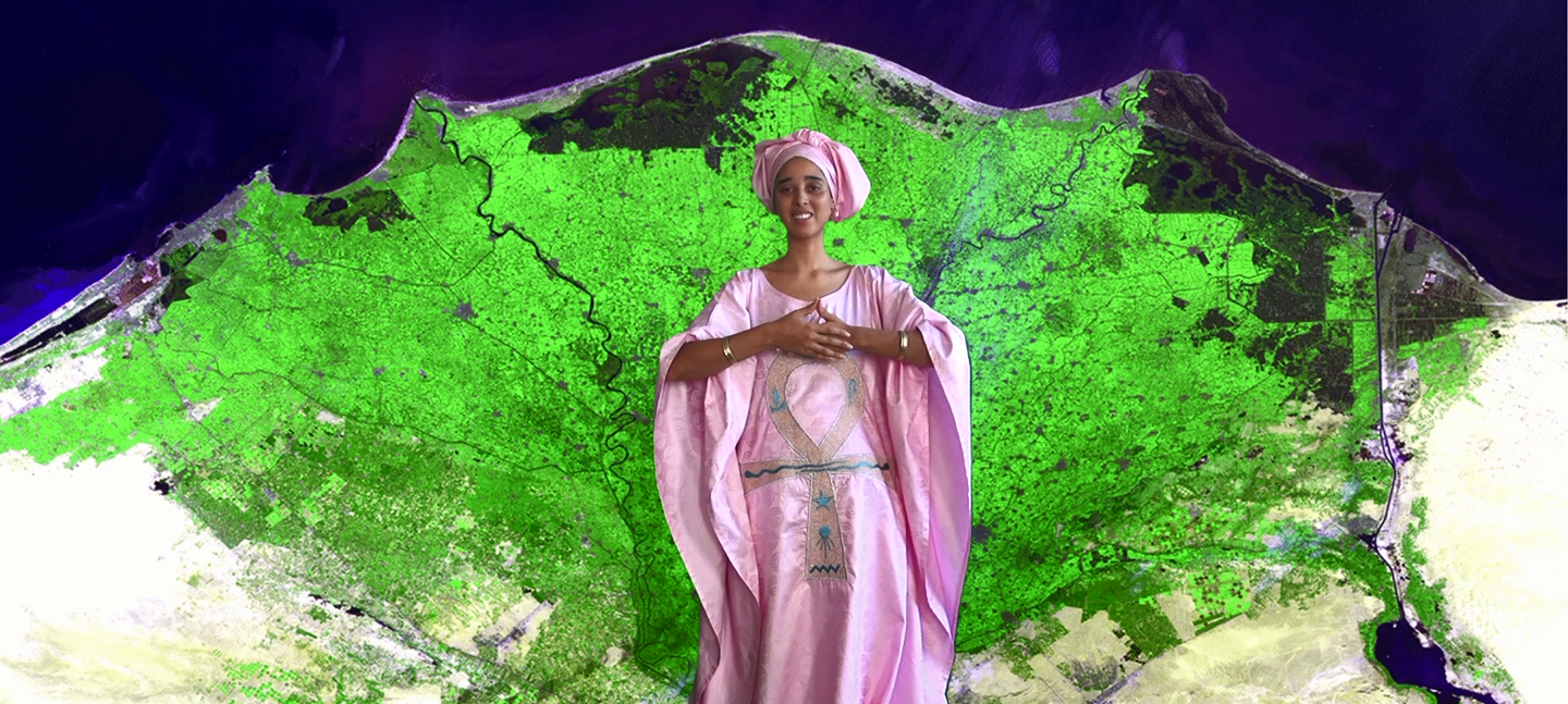 A still from Tabita Rezaire's MerKaBa for the Hoeteps, Tabita stands in a flowing pink, satin gown and head wrap, against a digital backdrop of a green aerial view of land mass and the deep blue of sky.