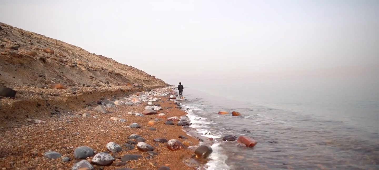 A still from a work by Dina Mimi. A small lone figure stands at the centre of shot. They are stood on a steep, pebble laden beach, with the sea on their right. It is overcast and the figure is turned away from the camera, and looks to be moving away.