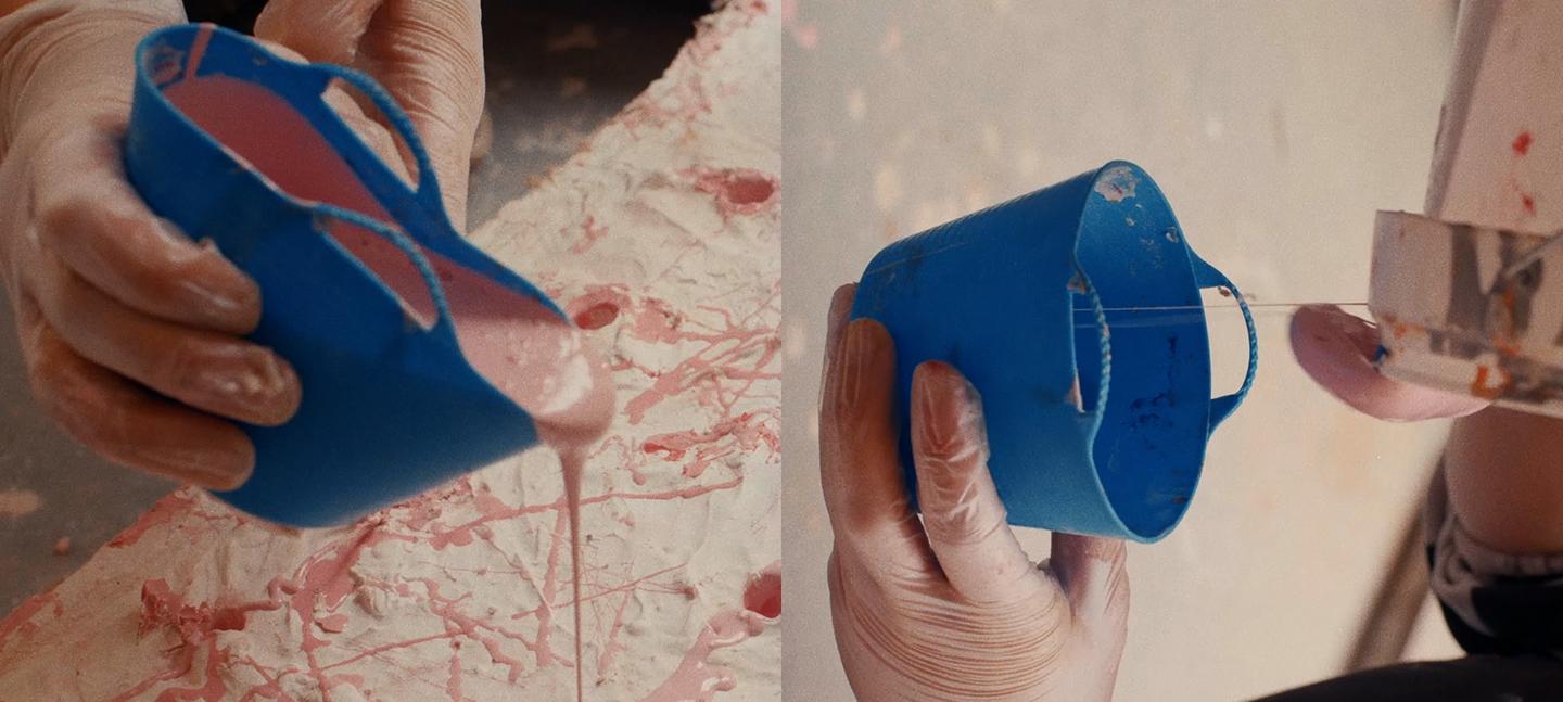 A still from the film Holly Hendry: The Art of Chaos. Gloved hands are shown pouring a pastel pink thick substance from a blue container into a textured white surface.