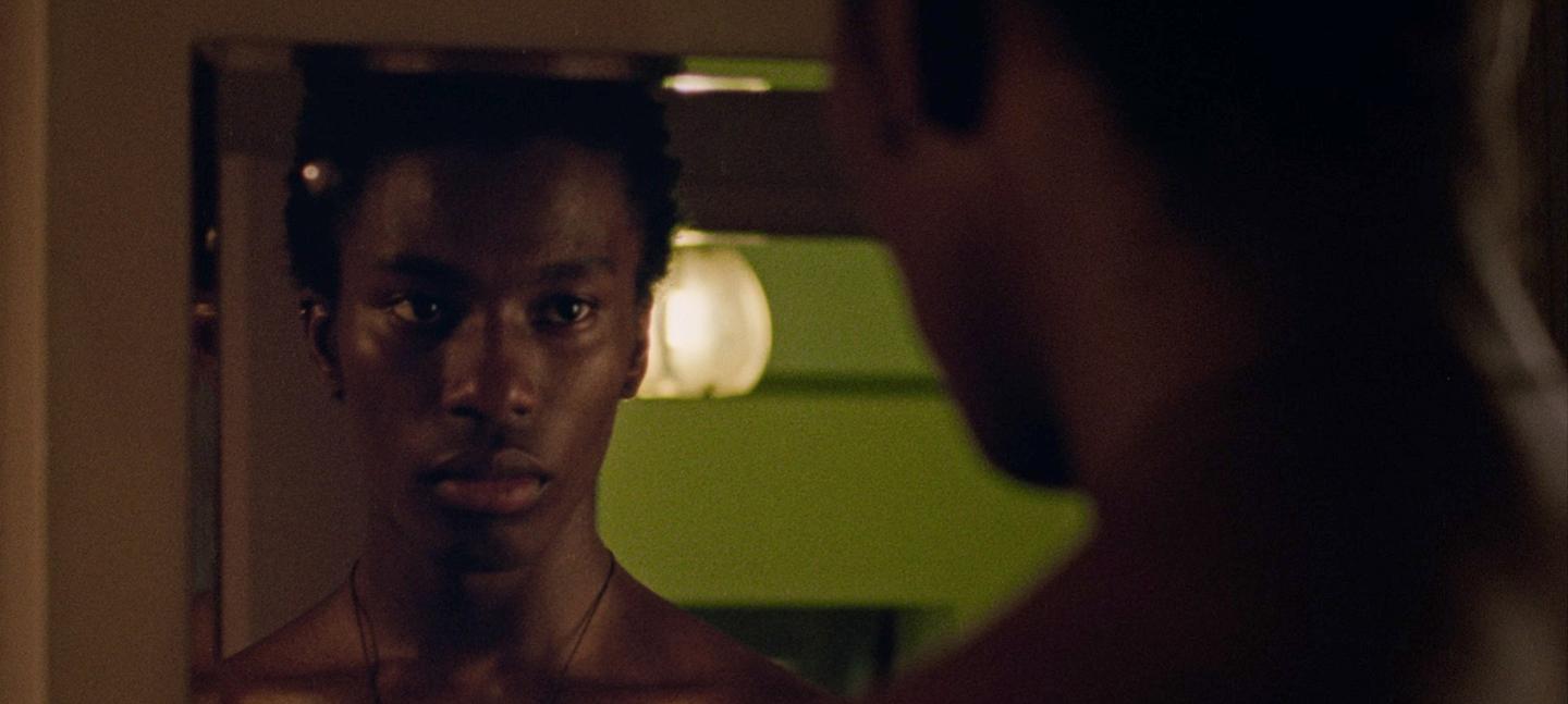 A still from a film by Iggy London. It shows a Black man, loking into a mirror, where we can see a reflection of his face.