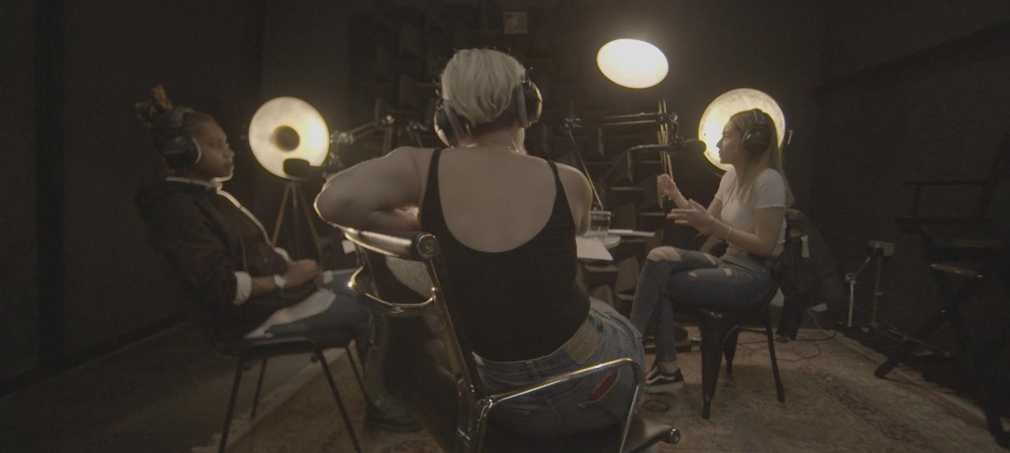 A photo of Jessie recording with two other speakers in a room with spotlights and recording equipment. 