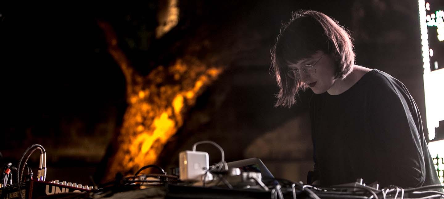 Joanne stands behind a table with a laptop and other electronic music equipment, performing.