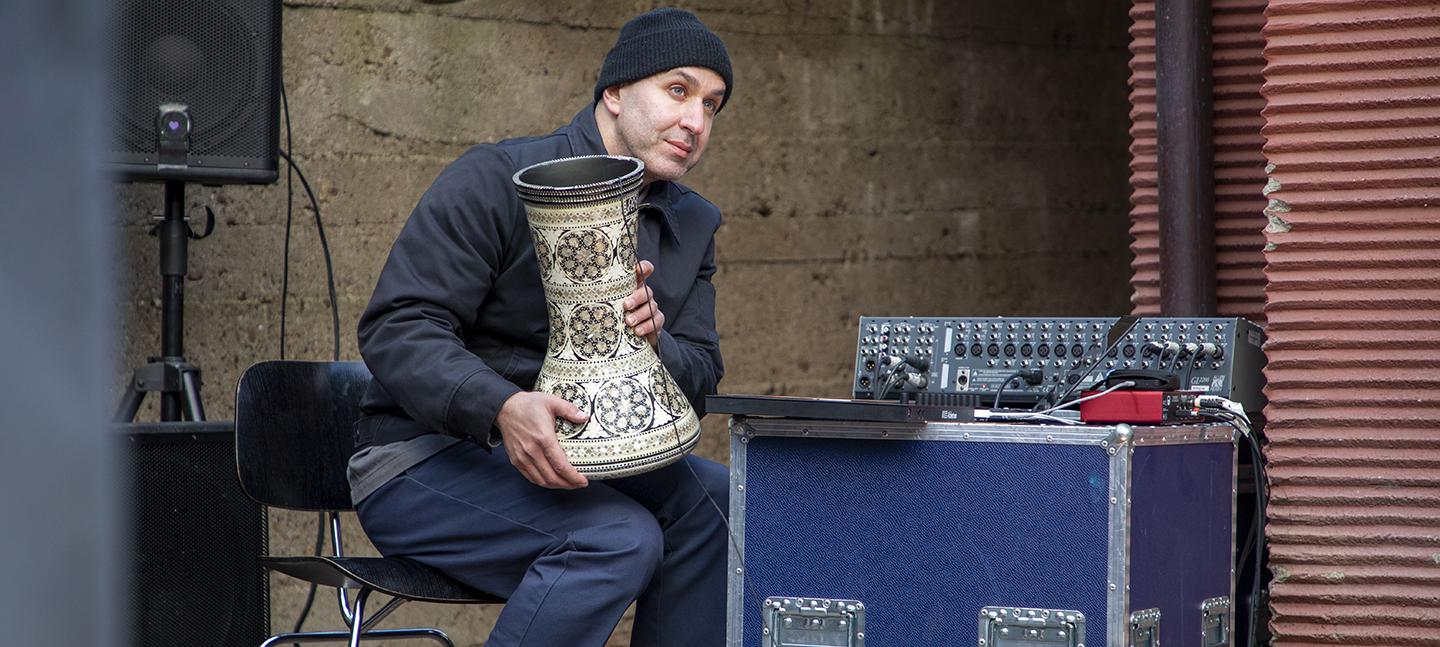 Documentation of Joe Namy's work at Intermissions in Portikus, Frankfurt. At an improvised performance a white man holds a drum in his lap. He sits on a chair next to a sound mixer resting upon a crate with wheels.