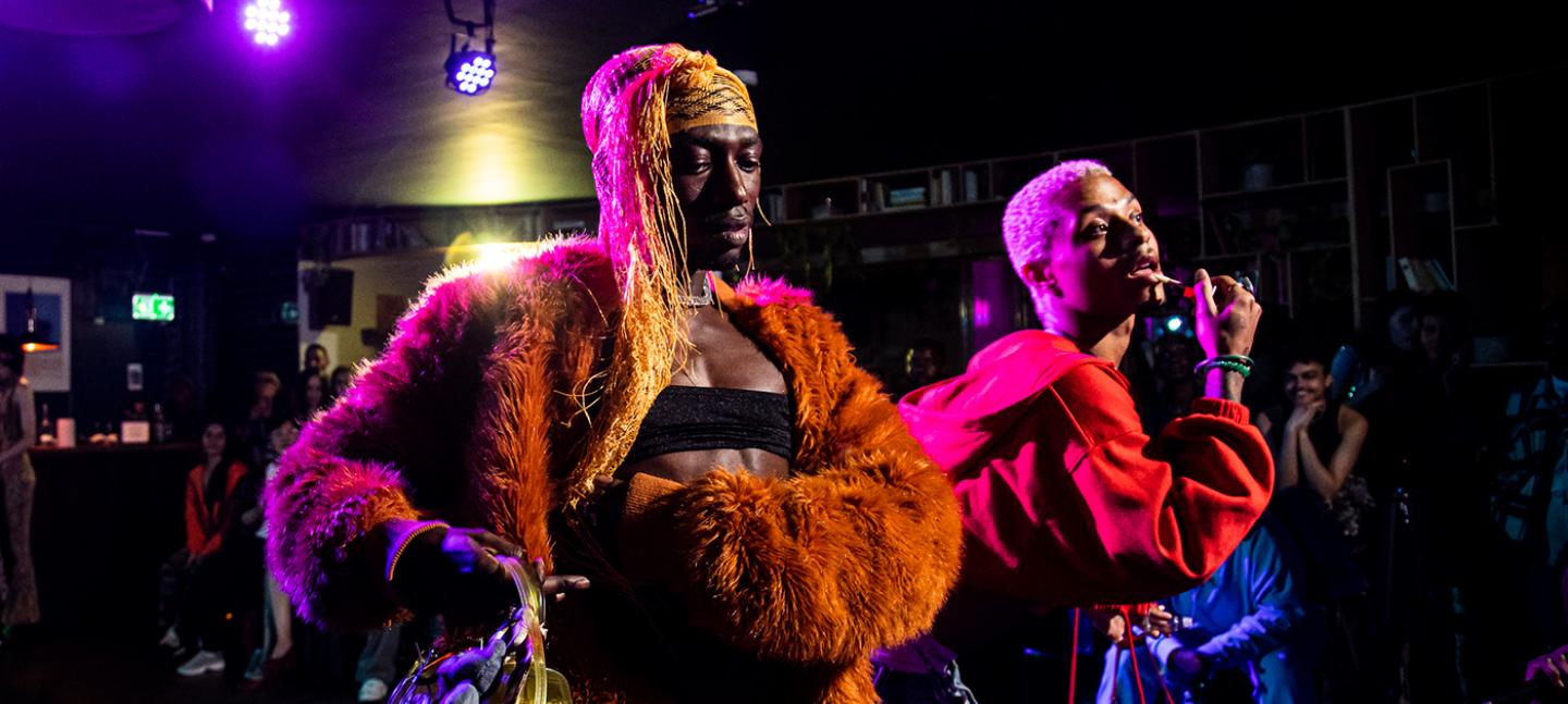 A photo of Vogue performers on the runway. One is wearing an orange fur jacket and a headscarf. The other is wearing a red jacket and applying lip gloss.