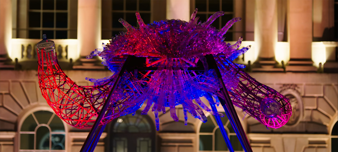 Close up of Leeroy New's installation The Arks of Gimokudan in the courtyard at Somerset House. It shows one of the sculptures, made up of reused plastic to look like a creature with arms, lit up by ambient lighting of pink, red and purple.