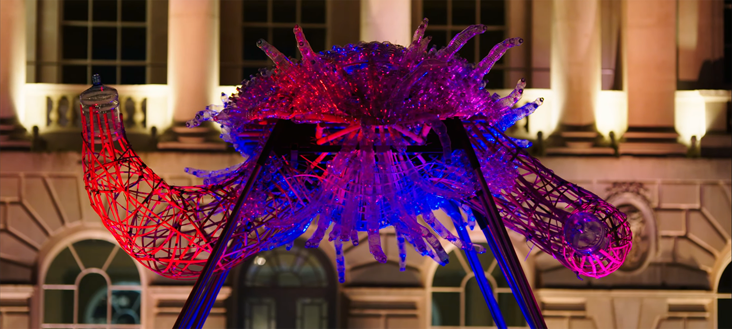 A photo of Leeroy New's installation The Arks of Gimokudan in the courtyard at Somerset House. It shows one of the sculptures, made up of reused plastic to look like a creature with arms, lit up by ambient lighting of pink, red and purple.