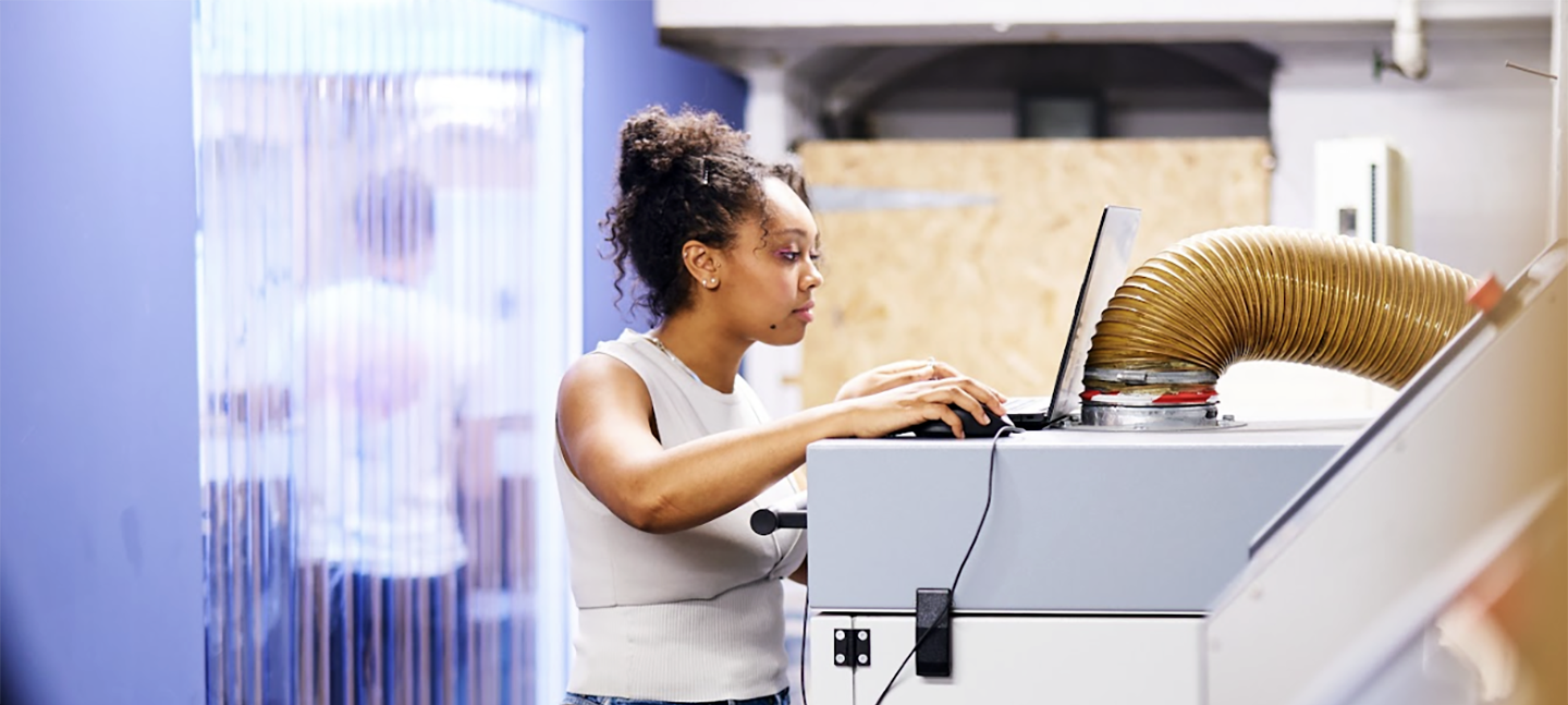 A young creative stands in front of a laptop in a workshop / studio space.