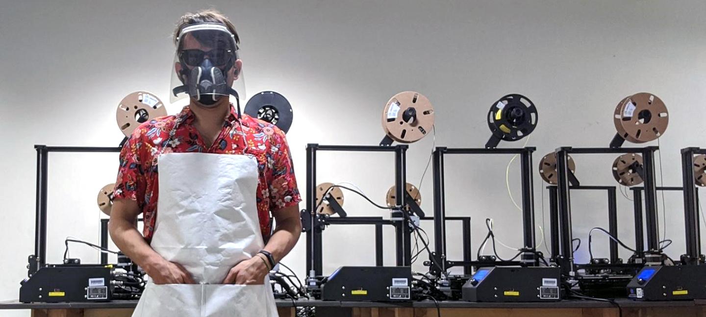 A photo of a man stood in front of 3-D printing equipment wearing a red shirt and a plastic face shield.