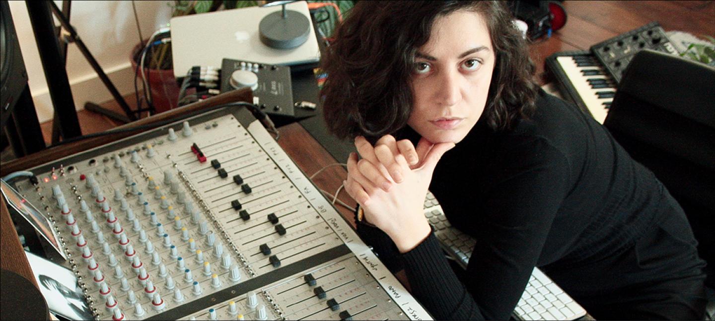 A photo of Marta Salogni. Marta is a white woman with short dark hair and brown eyes. Marta sit next to a soundboard with her head resting on clasped hands and looks up to the camera.