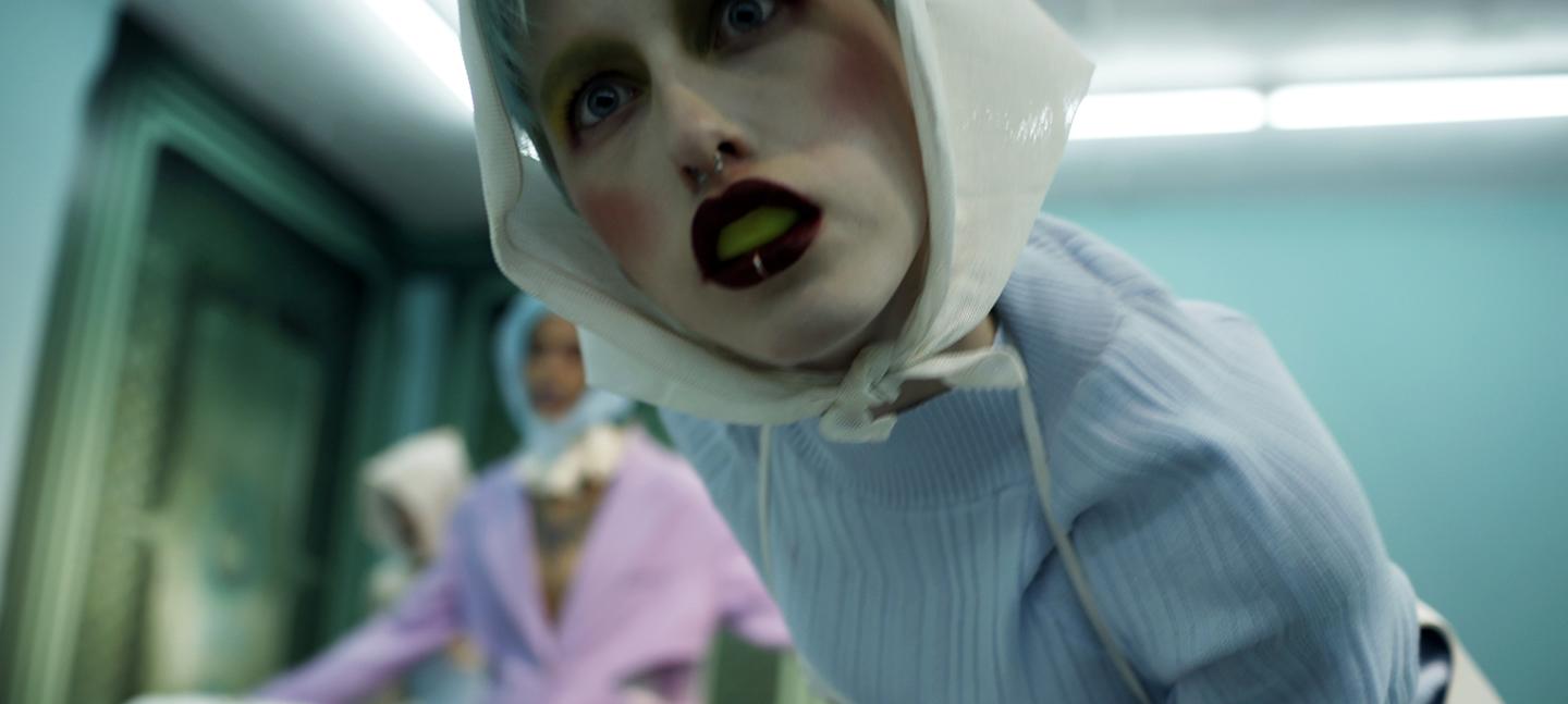 An installation image of Memeplex at Seventeen Gallery, where Omsk and Joey Holder both exhibited work. A woman leans over a camera positioned from a hospital bed. The women wears white powder and rouge, with dark burgundy lips and yellow inside her mouth