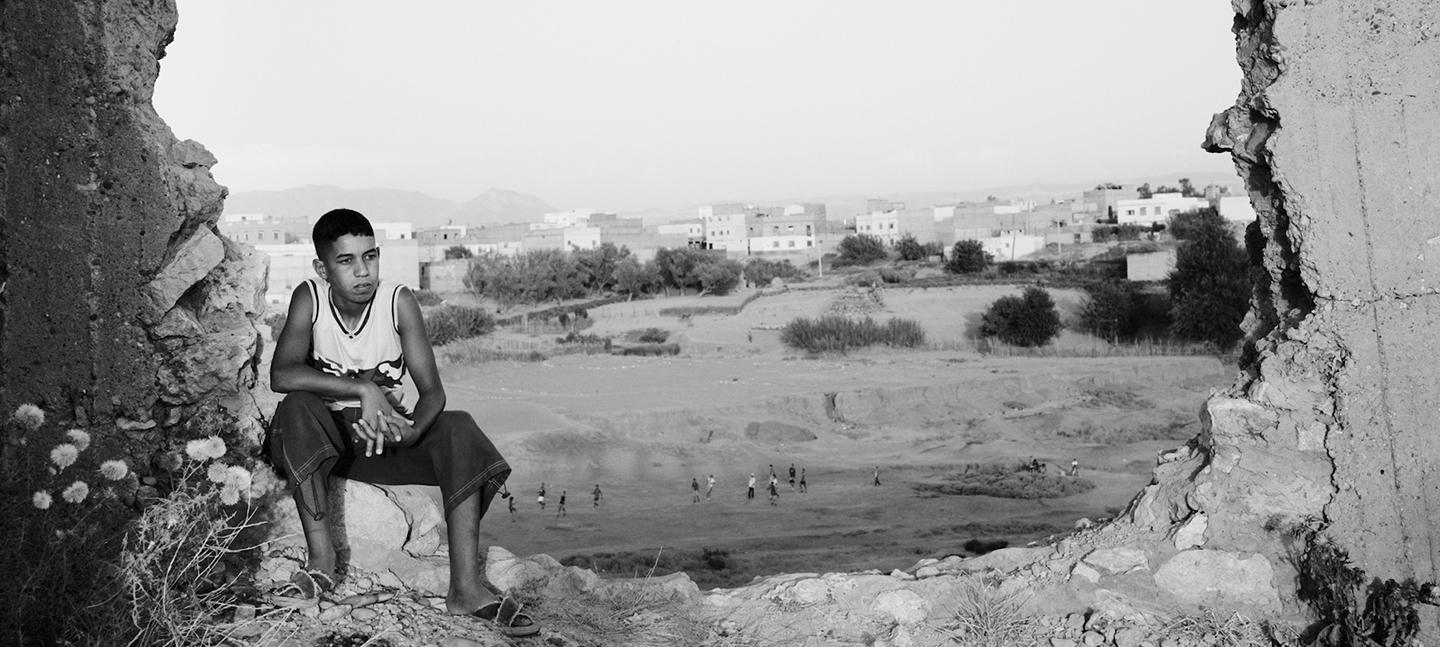 A black and white photo of a boy sitting on the broken remains of a wall that has been damaged and knocked down.