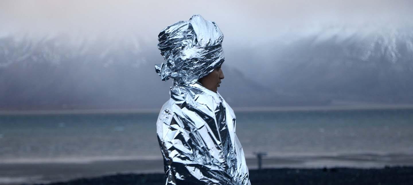An artwork. A woman stands, wrapped in a foil blankets, that cover her body and hair. In the background are snow-capped mountains and a body of cool blue water.
