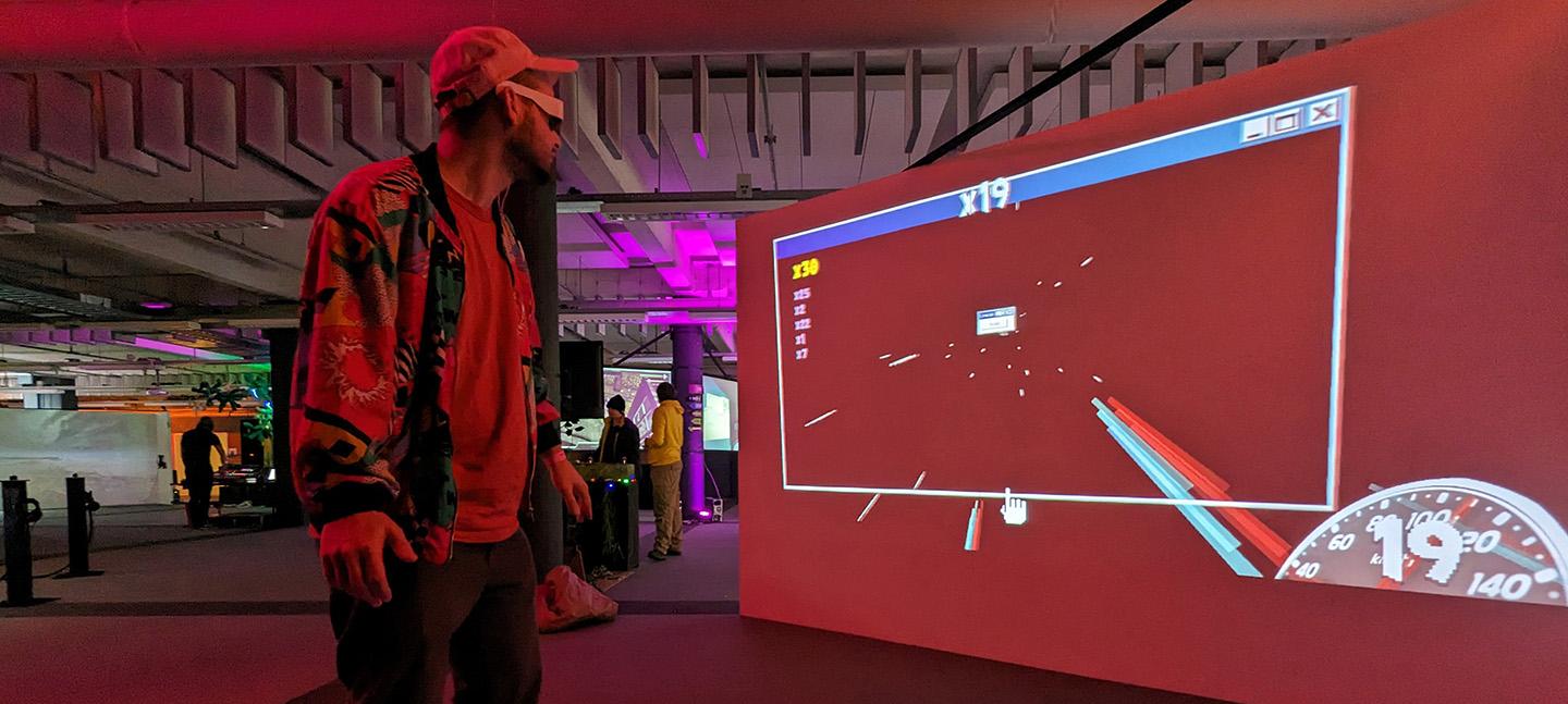 A man wearing a baseball cap, participating in a game, wears VR goggles looks at a large screen that simulates the experience of flying through space. On the right corner of the screen is a speedometer.