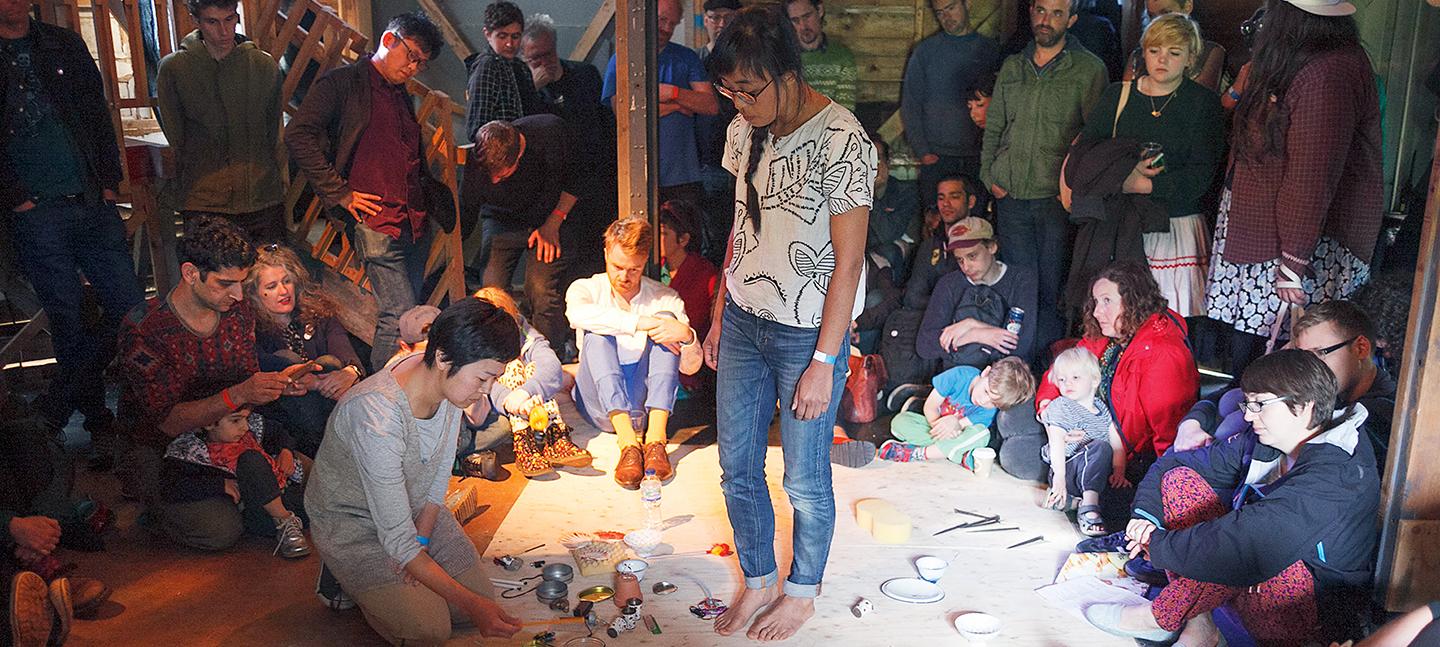 A photo of O YAMA O performing. A pair of slender young people, one kneeling, the other stood, are both atop a big sheet of paper, with bowls, cups and plates scattered between them. They are surrounded by a crowd of people who are watching on.