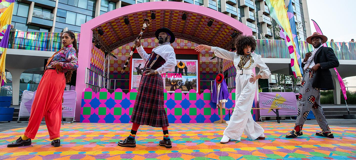 A photo of Patrick Ziza performing 'Dance by Design'. Four performers in Dandy clothing perform on stage. They are dressed in colourful shirts, hats and suit trousers, pulling suave poses.