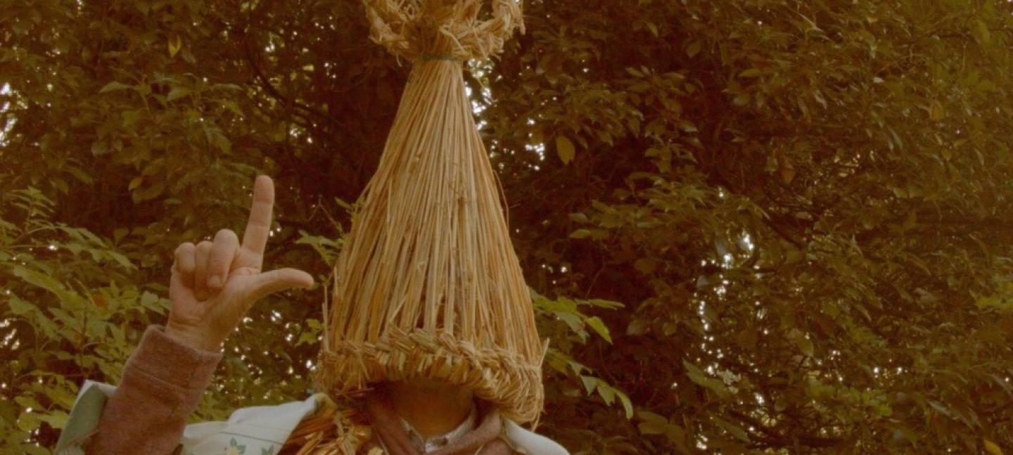 A photo of a man with a straw head piece that covers his face. The right hand is held up with only the forefinger and thumb showing.