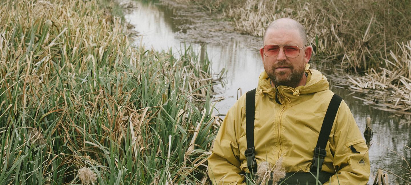 Raf Rundell wears a mustard yellow rain coat and waders and stands in tall reeds by a stream.
