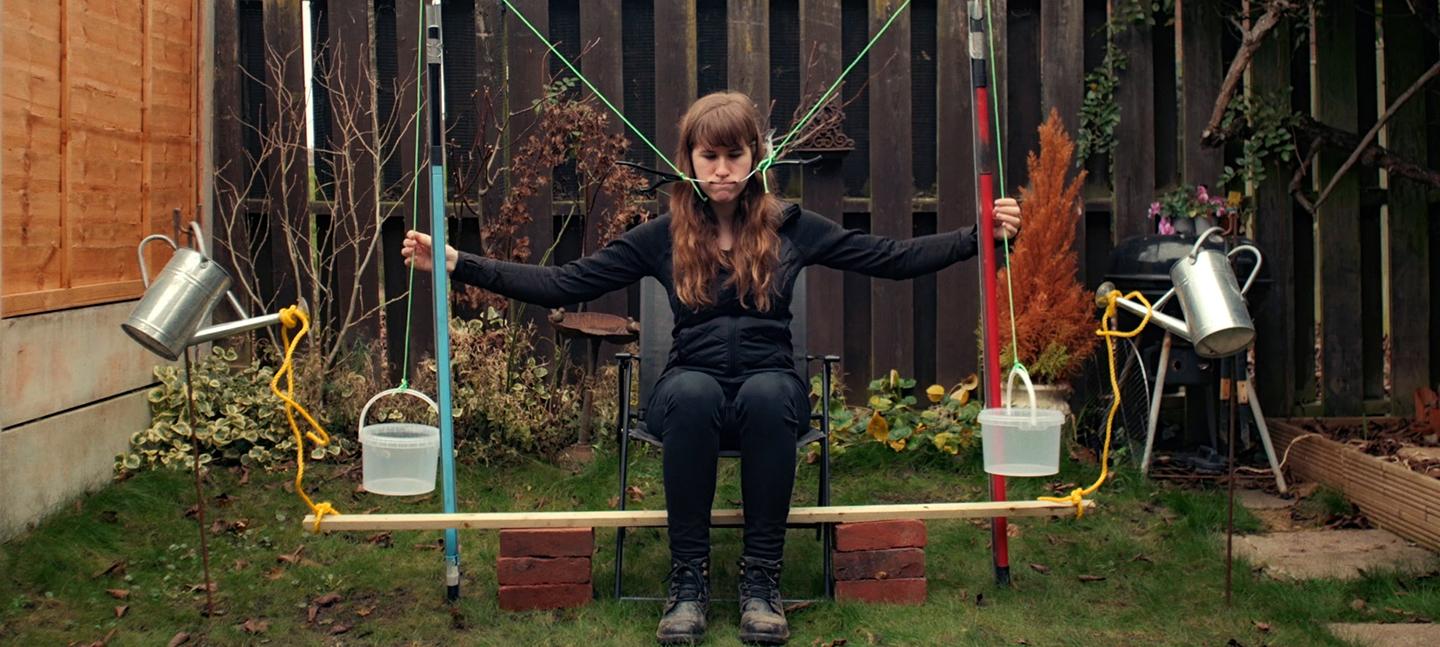 A photo of Rebecca Moss operating her smile machine. She sits in a garden, with the hooks of two coathangers hooked around the inside of her cheeks. As she pulls on ropes the coat hanger hooks pull her mouth into a smile.