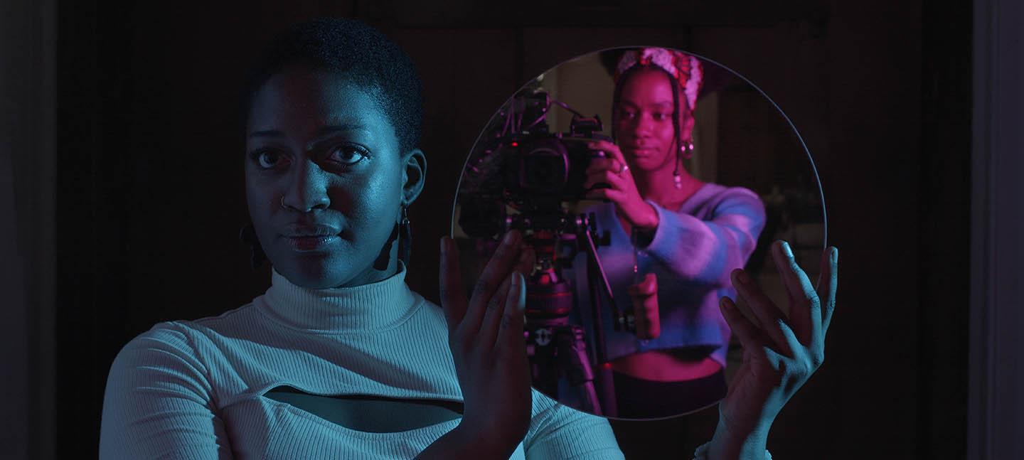 A still from the Future Producers' film Rising: A Manifesto. A Future Producer, Kayleigh, holds a circular mirror up to the camera. In the reflection you can see another Future Producer, Jahnavi, operating the camera capturing the image. 