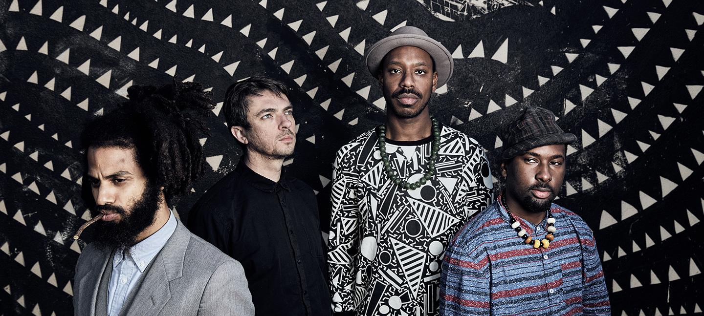 A photo of the band Sons of Kemet. Four men, stood against a patterned black and white backdrop.