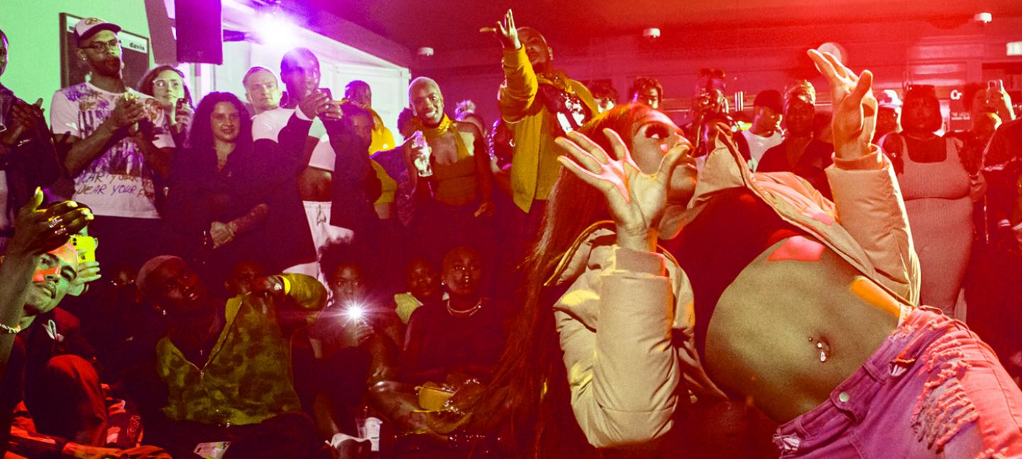 A photo of a Vogue Ball. In the foreground a performer is in motion, throwing their hands up. In the background onlookers cheer.