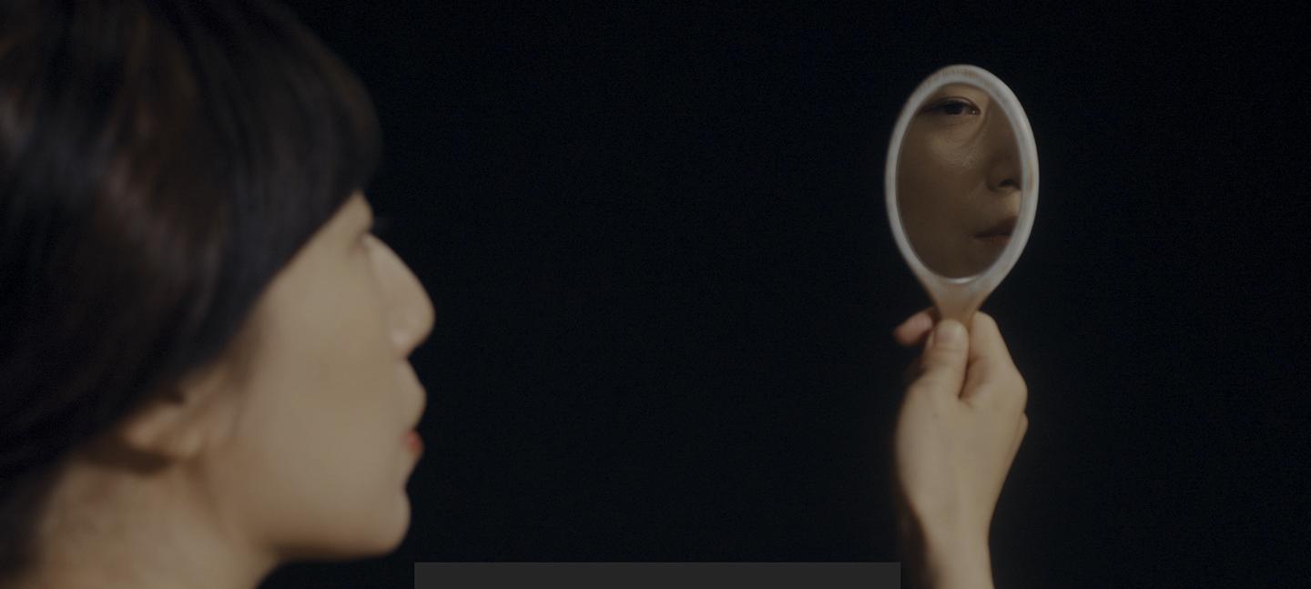 A still from a film by Shen Xin. A person holds a vanity mirror up - from it you can see the person's face from another angle.
