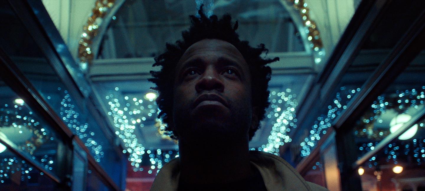 A still from the film Simeon Barclay: Defying the Norm. In it the artist Simeon Barclay, a Black man with medium length hair looks upwards as he stands in a shopping arcade in Leeds, that is lit with fairy lights in the darkness.