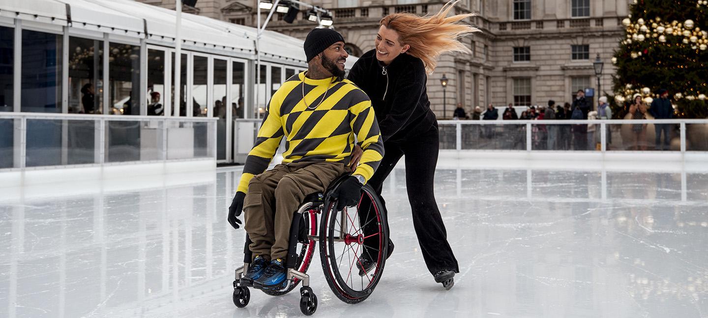 A wheelchair user and their partner are on the ice at Somerset House, smiling at one another as they speed past on the ice.
