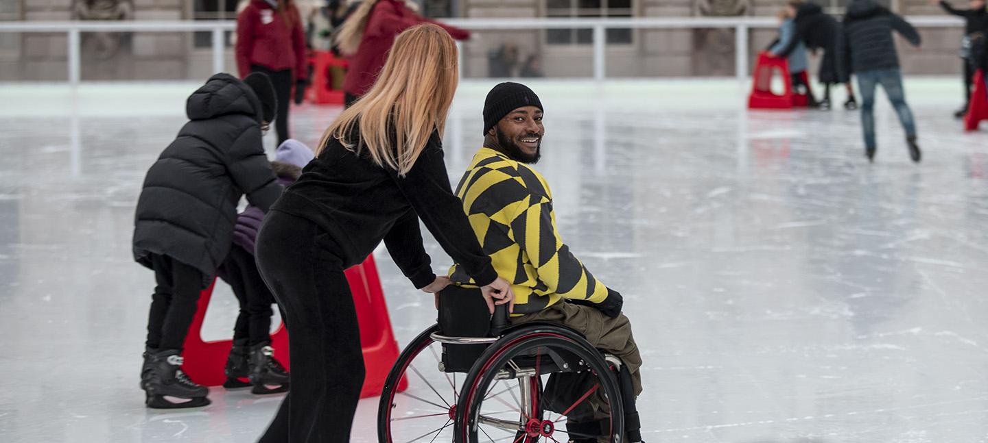 Revellers on the ice at Somerset House. One man, wearing a black and yellow jumper sits in a wheelchair looking behind him.