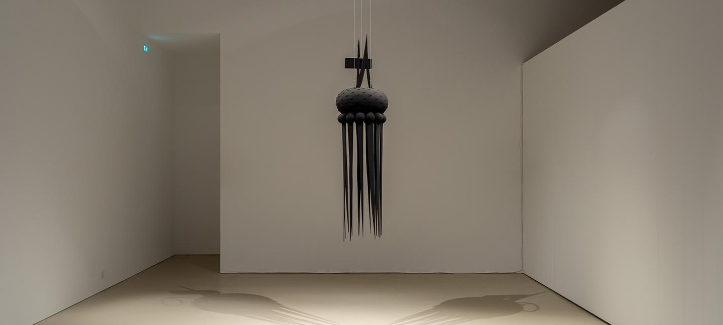 A photo of an installation artwork by Sonya Dyer. It depicts a black sculpture hanging from the ceiling in a white room.