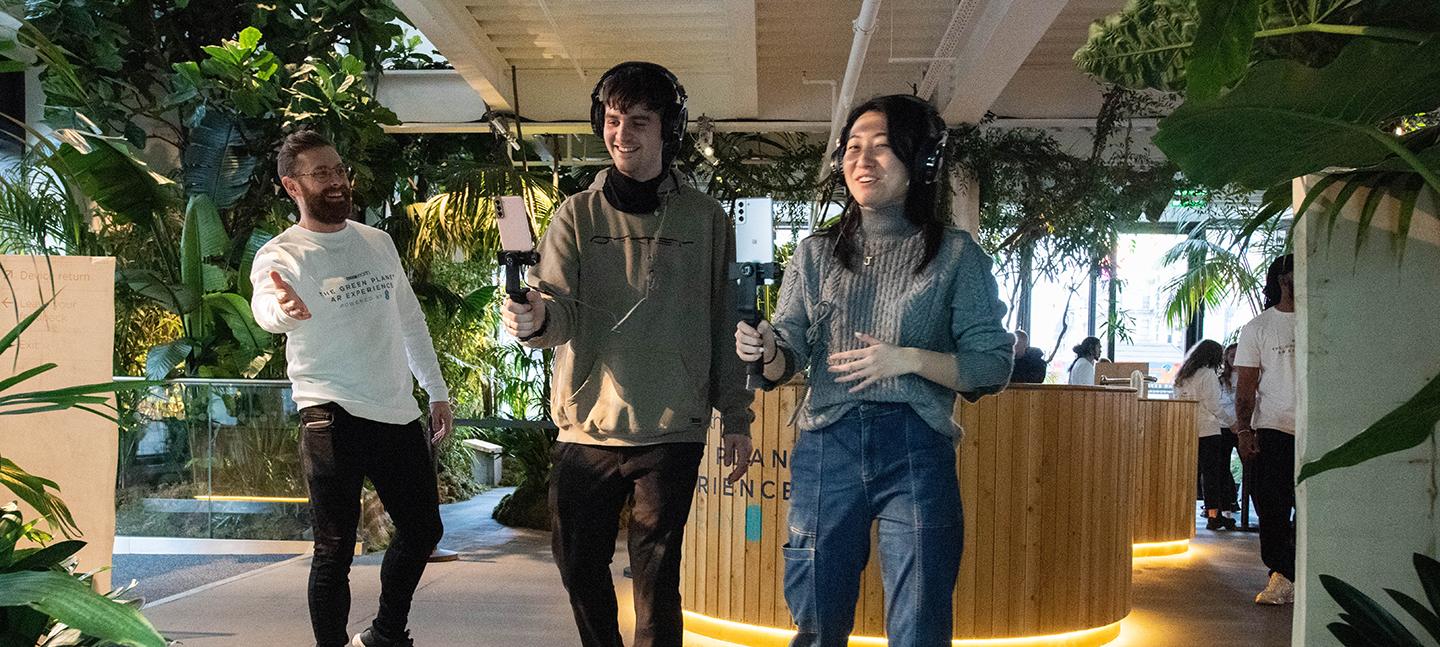 A photo of placement participants at work on 'Green Planet'. Two young people hold technical film equipment out in front of them, while someone else looks on with their arms thrown wide. There are green plants all around them.